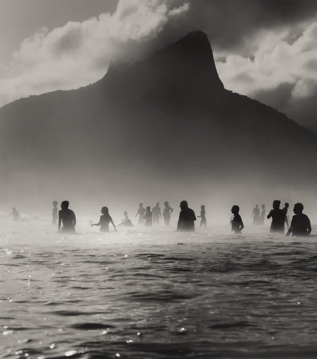 Black and white photograph of silhouetted figures standing in calm water beneath a dramatic mountain peak